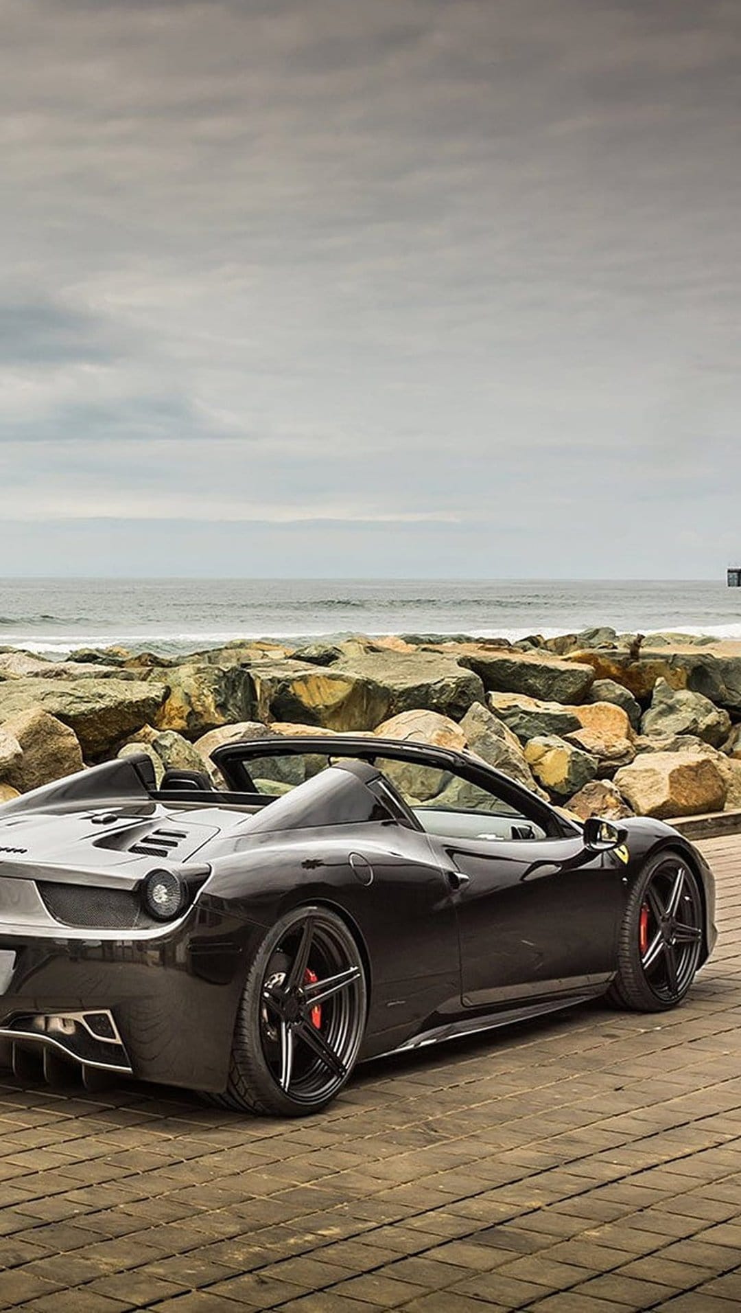 Black Ferrari convertible parked on a wooden deck overlooking a rocky coastline and a misty grey ocean horizon.