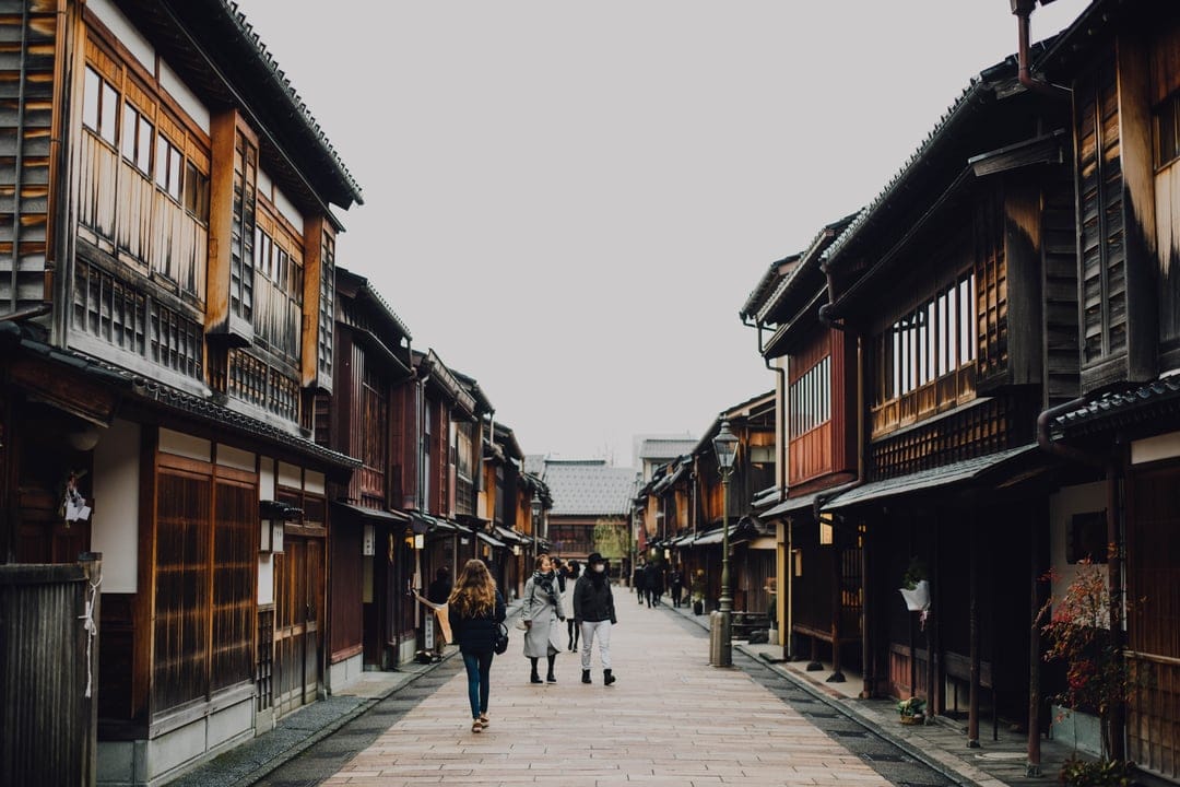 Historic wooden buildings line a stone-paved street with walking figures under a soft overcast sky.