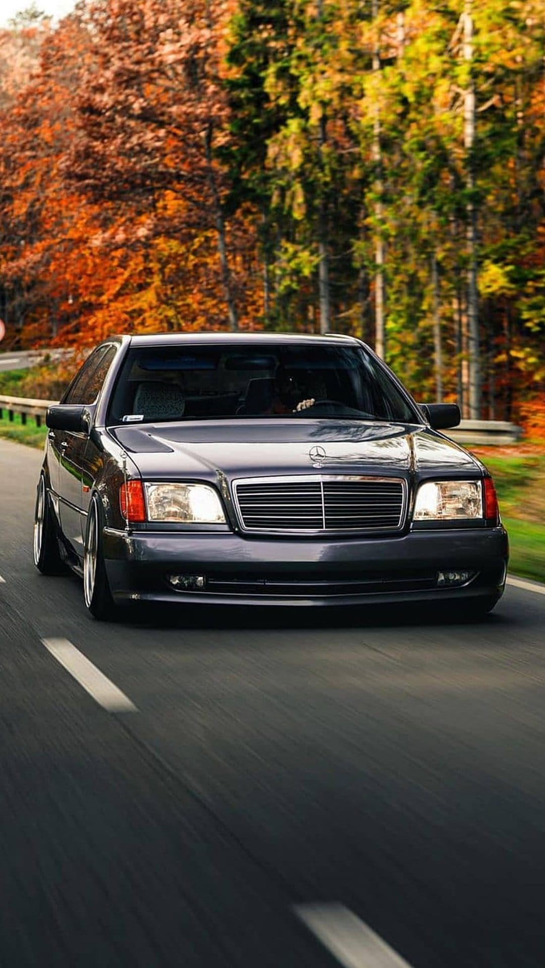 Dark Mercedes W140 sedan drives quickly through a forest of orange and yellow autumn trees with headlights on.