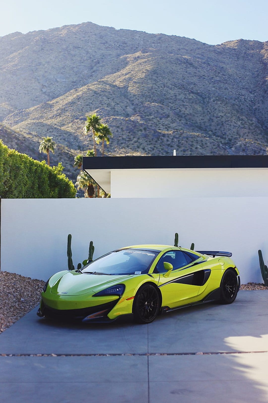 Lime green McLaren sports car parked on a white driveway beside cacti and palms under a clear mountain sky.