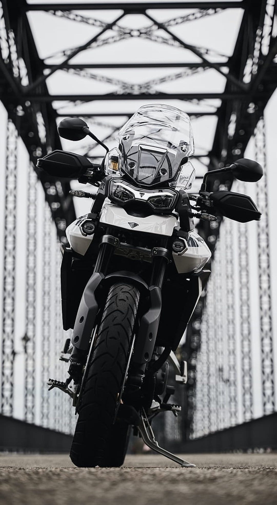 Triumph Tiger adventure motorcycle parked on a concrete road between the metal trusses of a large steel bridge.
