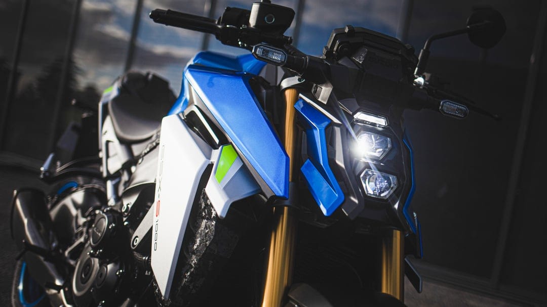 Suzuki GSX-S1000 sportbike with glowing LED headlights, blue and white fairings, and gold front forks.
