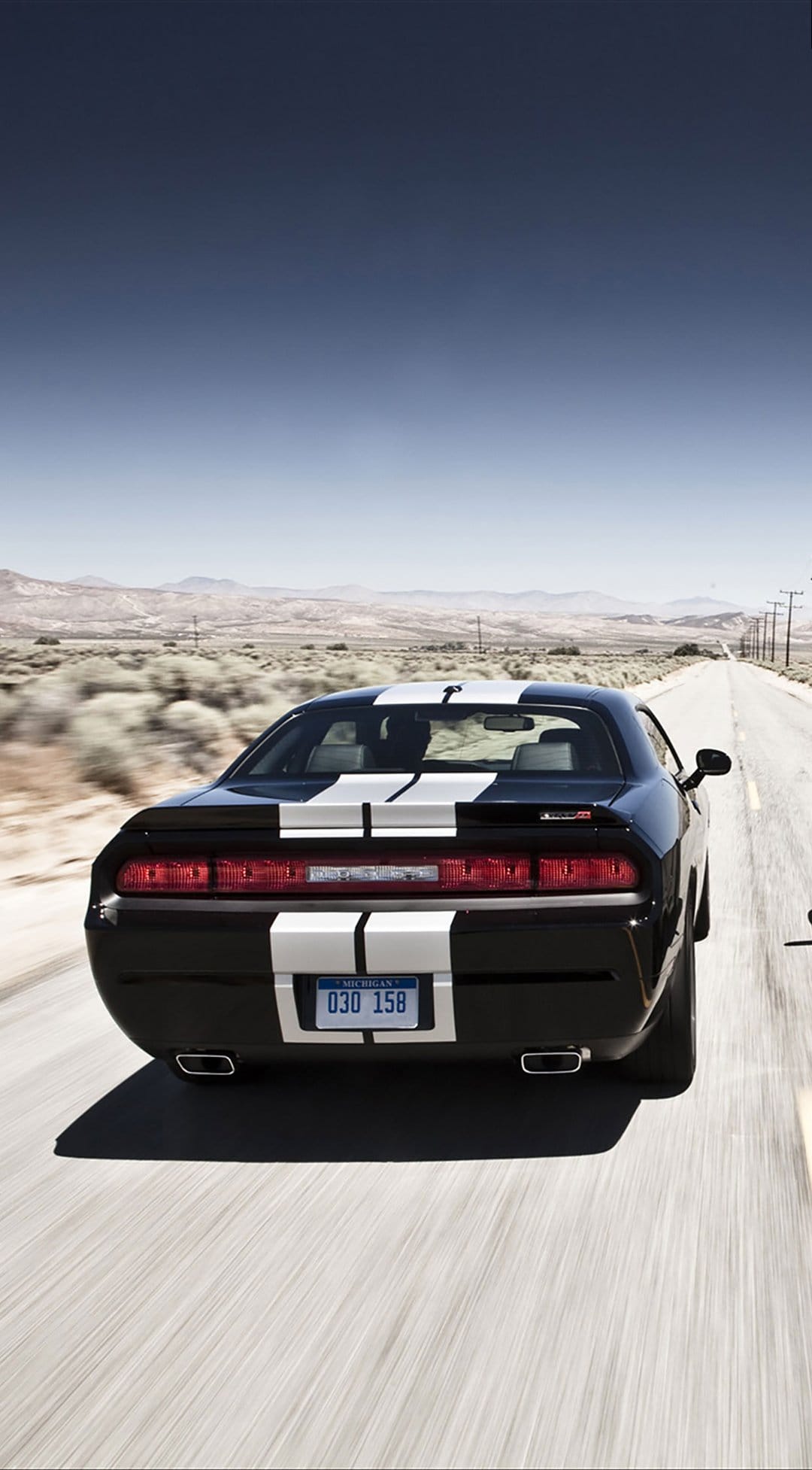 Black Dodge Challenger with white stripes driving on a straight desert highway under a clear blue sky.