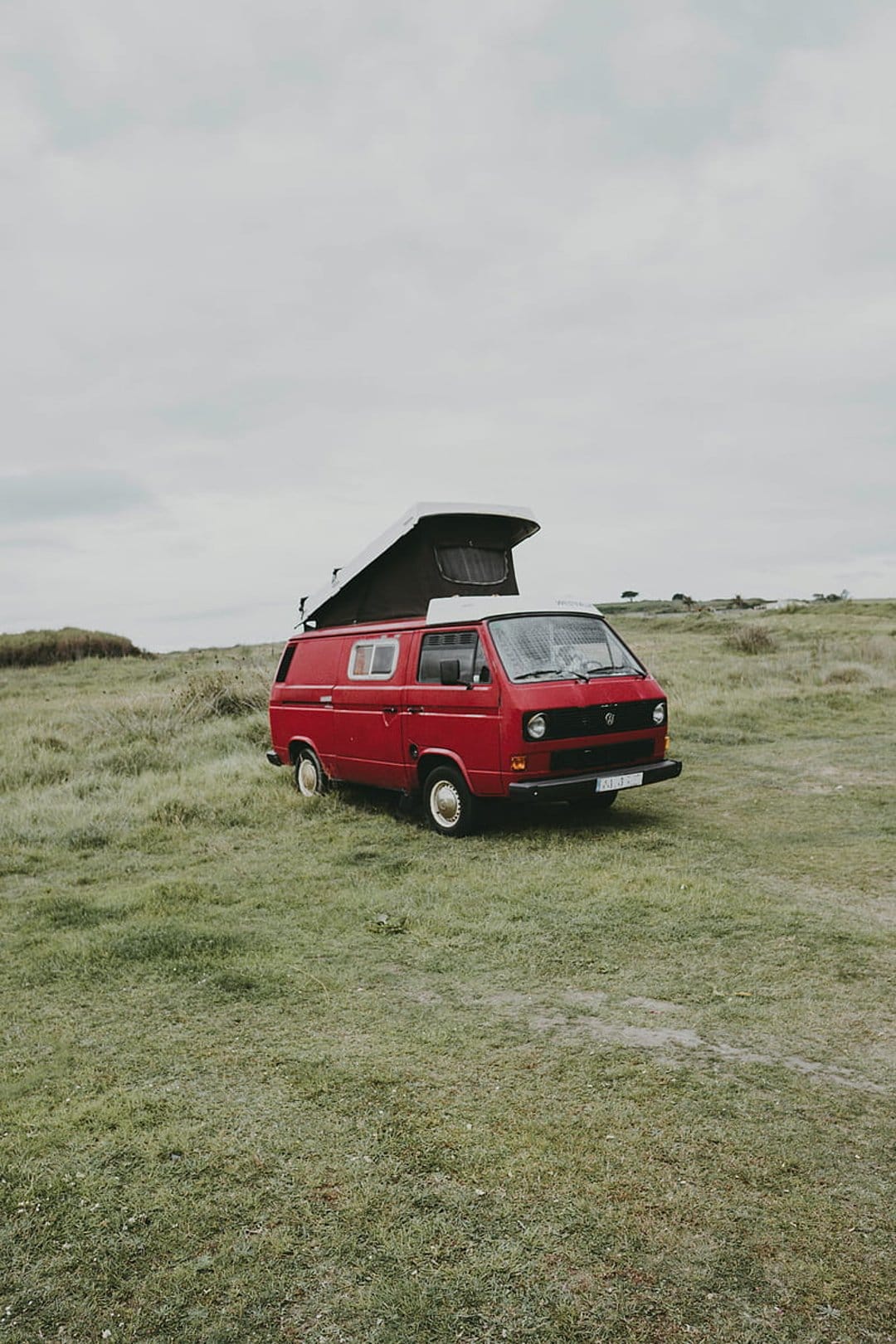 Red vintage camper van with a pop-top roof parked in a vast green field under a cloudy, overcast sky.