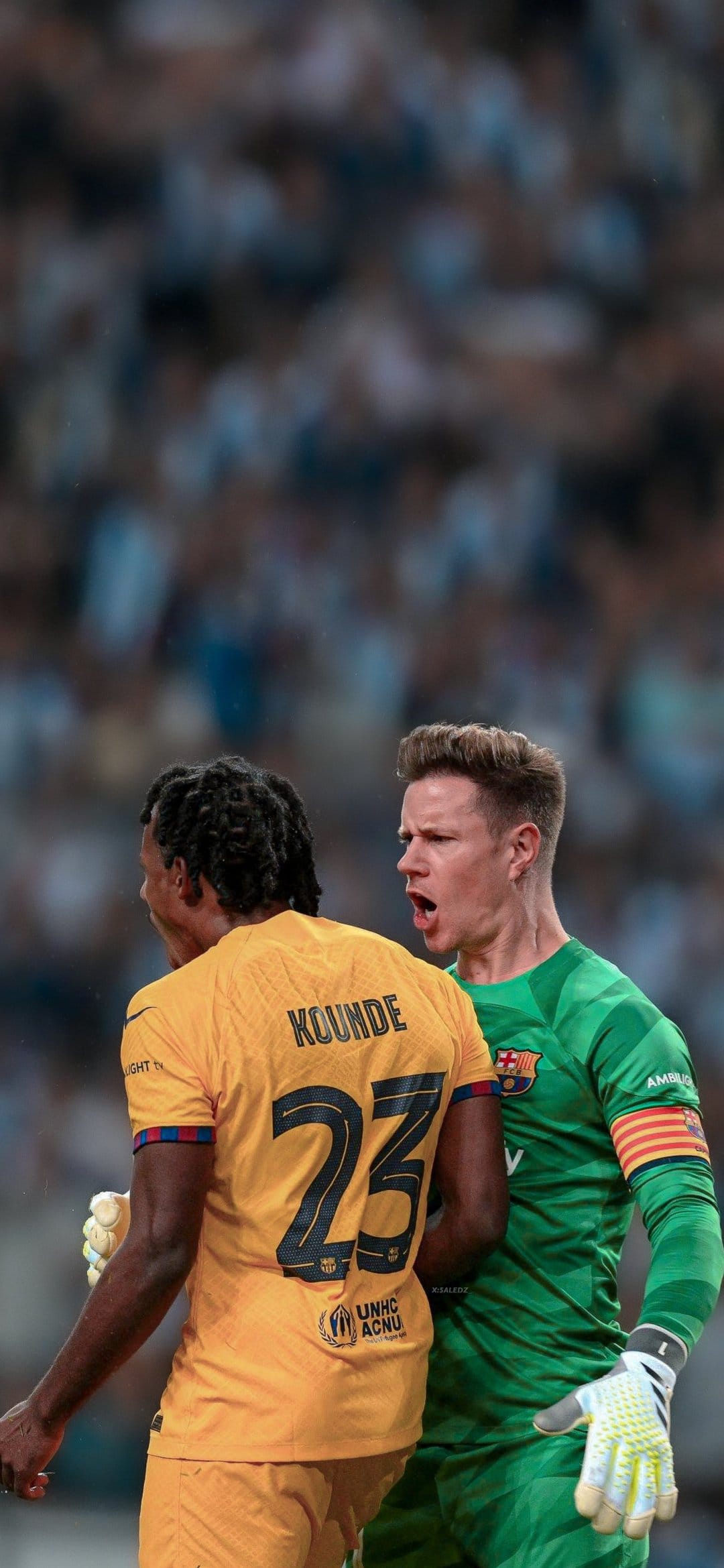 Jules Kounde in a yellow kit speaks with Ter Stegen in green during a professional soccer match on green grass.