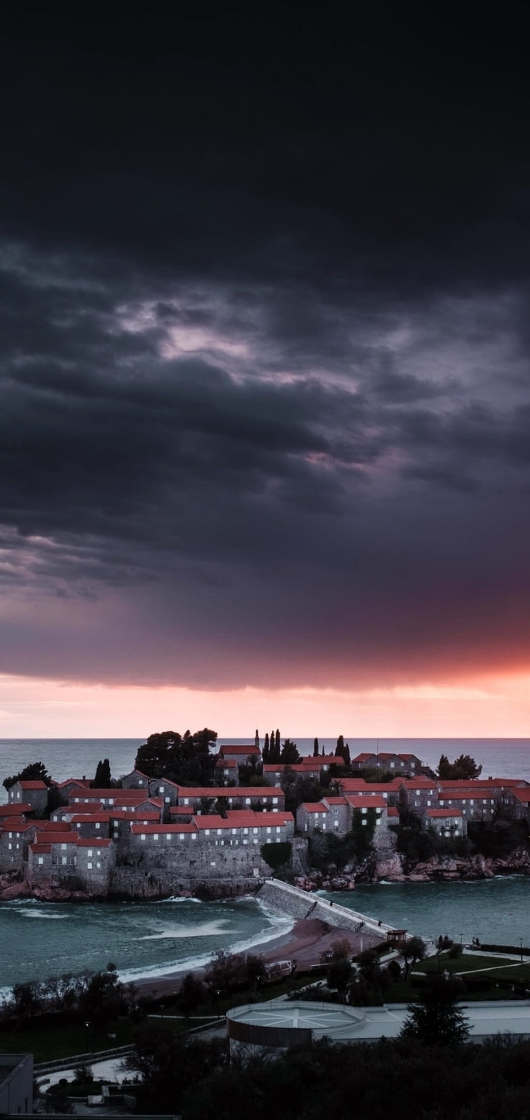 Mediterranean island village with red roofs and a stone causeway under a stormy purple and orange twilight sky.