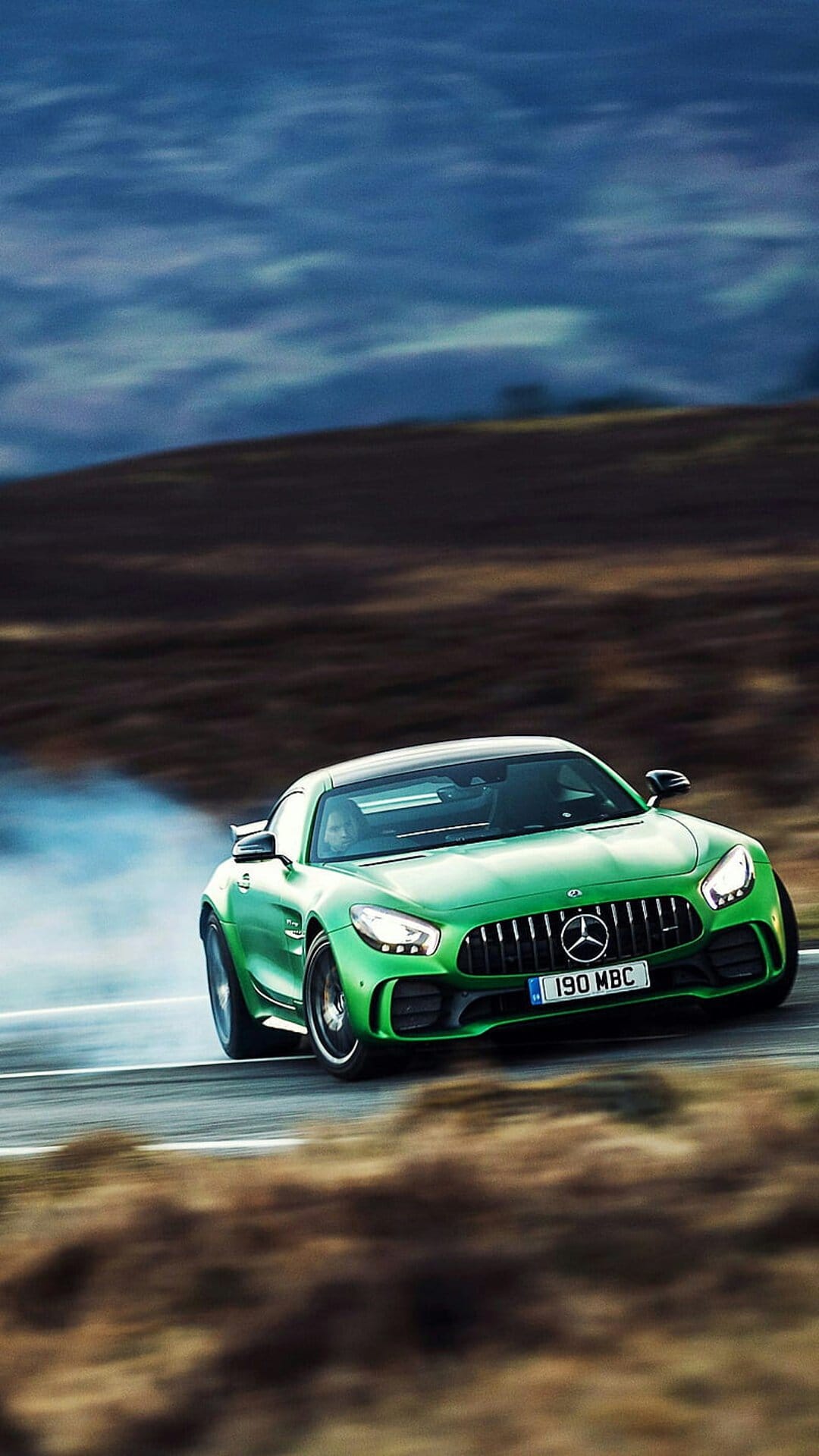 Green Mercedes-AMG GT car drifts sideways on a mountain road with white tire smoke against rolling hills.