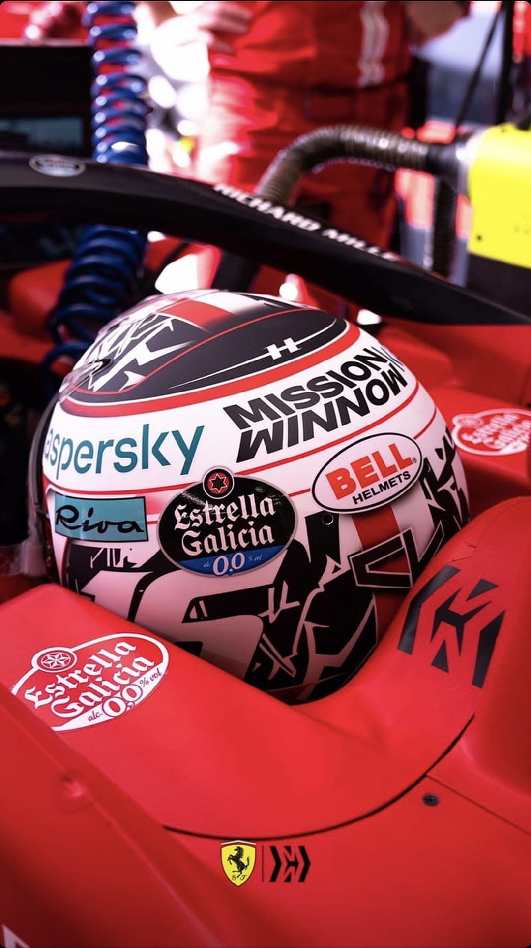 Red Ferrari Formula 1 racing helmet and steering wheel detail inside a carbon fiber cockpit with sponsor logos.