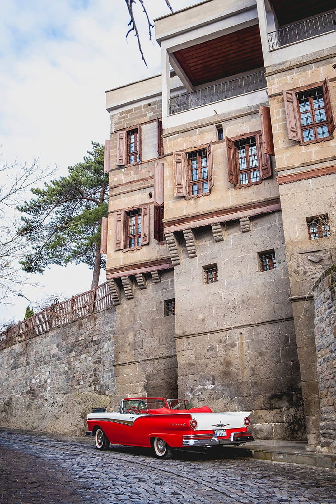 Red 1950s convertible parked on a cobblestone street in front of a historic stone building with wooden shutters.