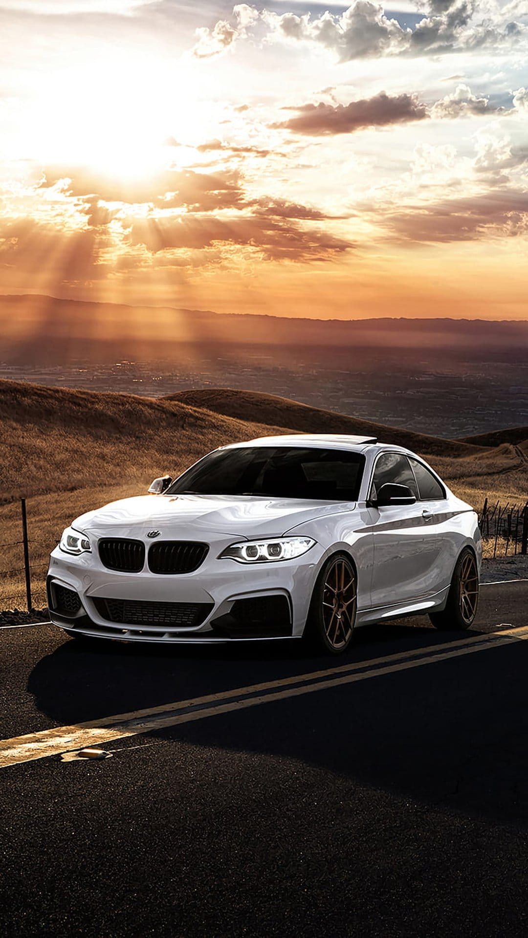 White BMW coupe maneuvers a winding mountain road under a golden sunset with radiant light beams through clouds.