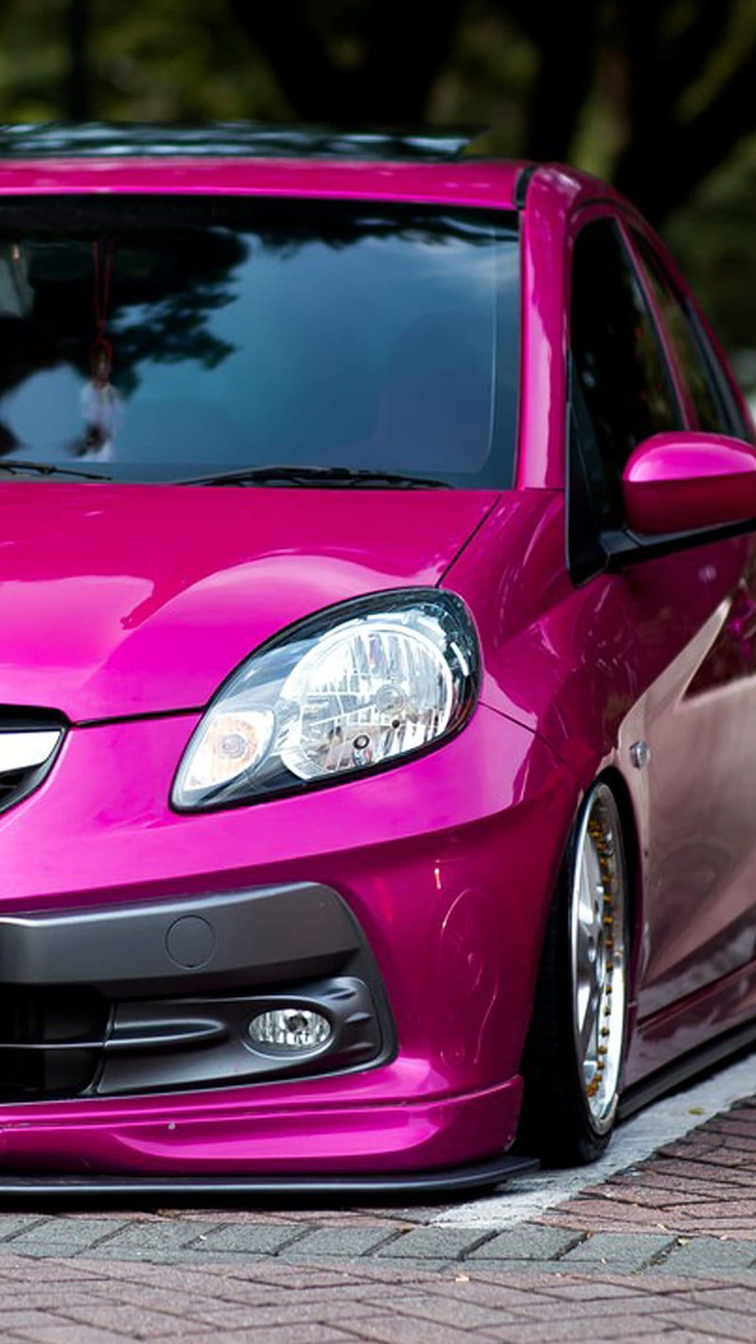Metallic pink car with gold rims and tinted windows parked on a brick road under soft daylight reflections.