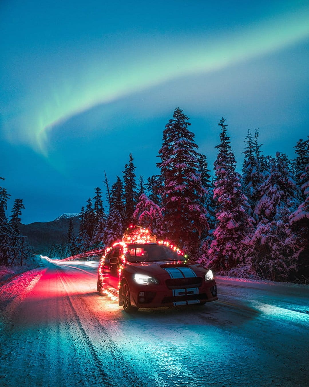 Red car with Christmas lights driving on a snowy road through pine trees under a green aurora borealis night sky.