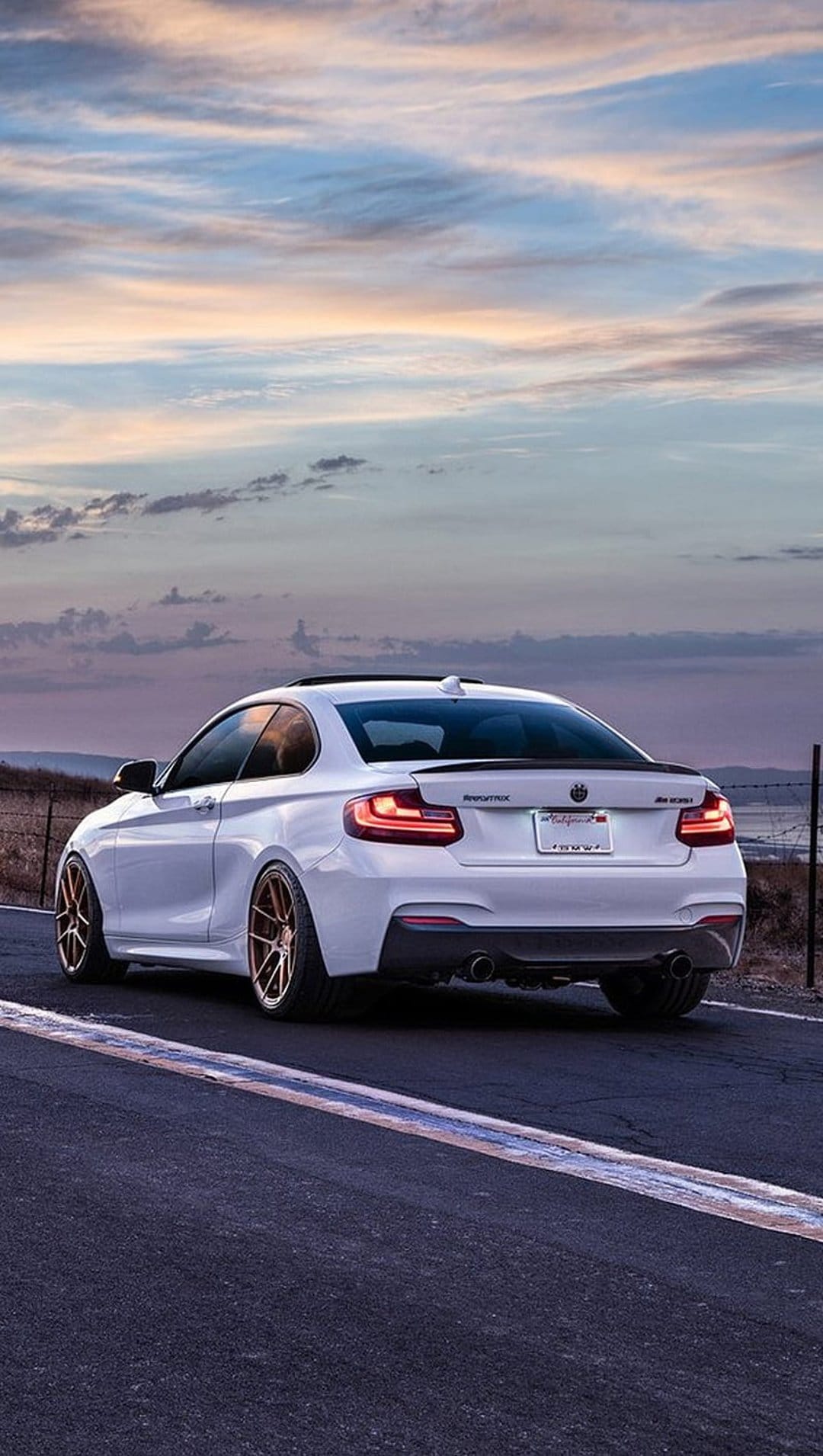 White BMW M2 with gold wheels parked on a road at sunset, featuring a rear spoiler and dramatic evening sky.