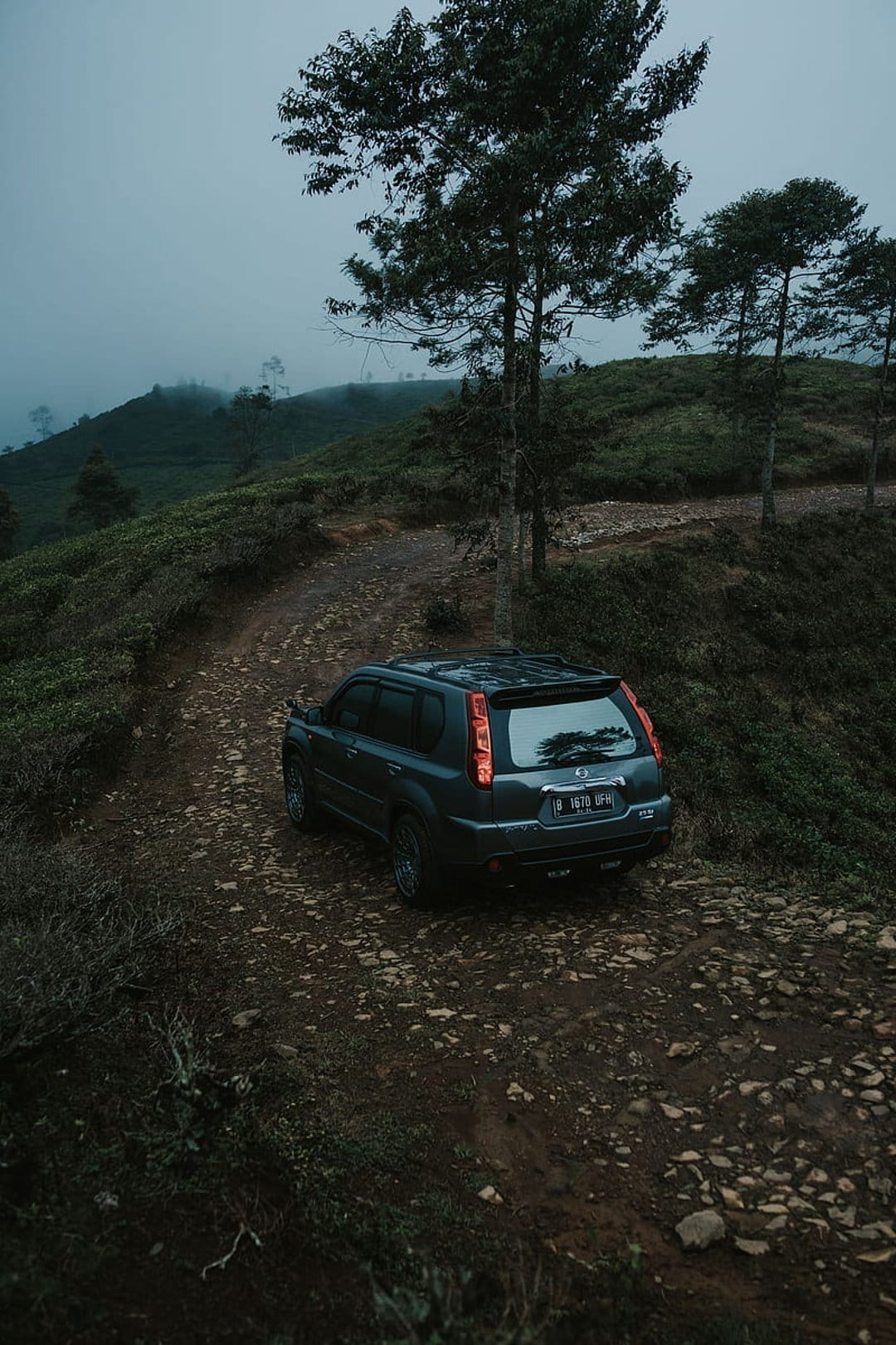 Dark SUV maneuvers along a muddy mountain road through a dense forest shrouded in thick morning mist and fog.
