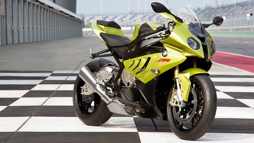Acid green BMW S1000RR sportbike parked on a checkered racetrack with paddock buildings and a clear sky background.