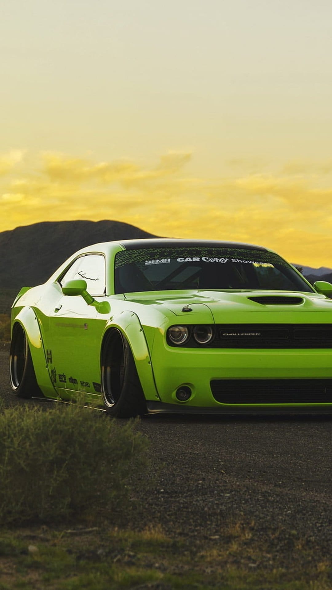 Lime green Dodge Challenger widebody on a gravel road under a yellow sunset sky with distant rolling hills.