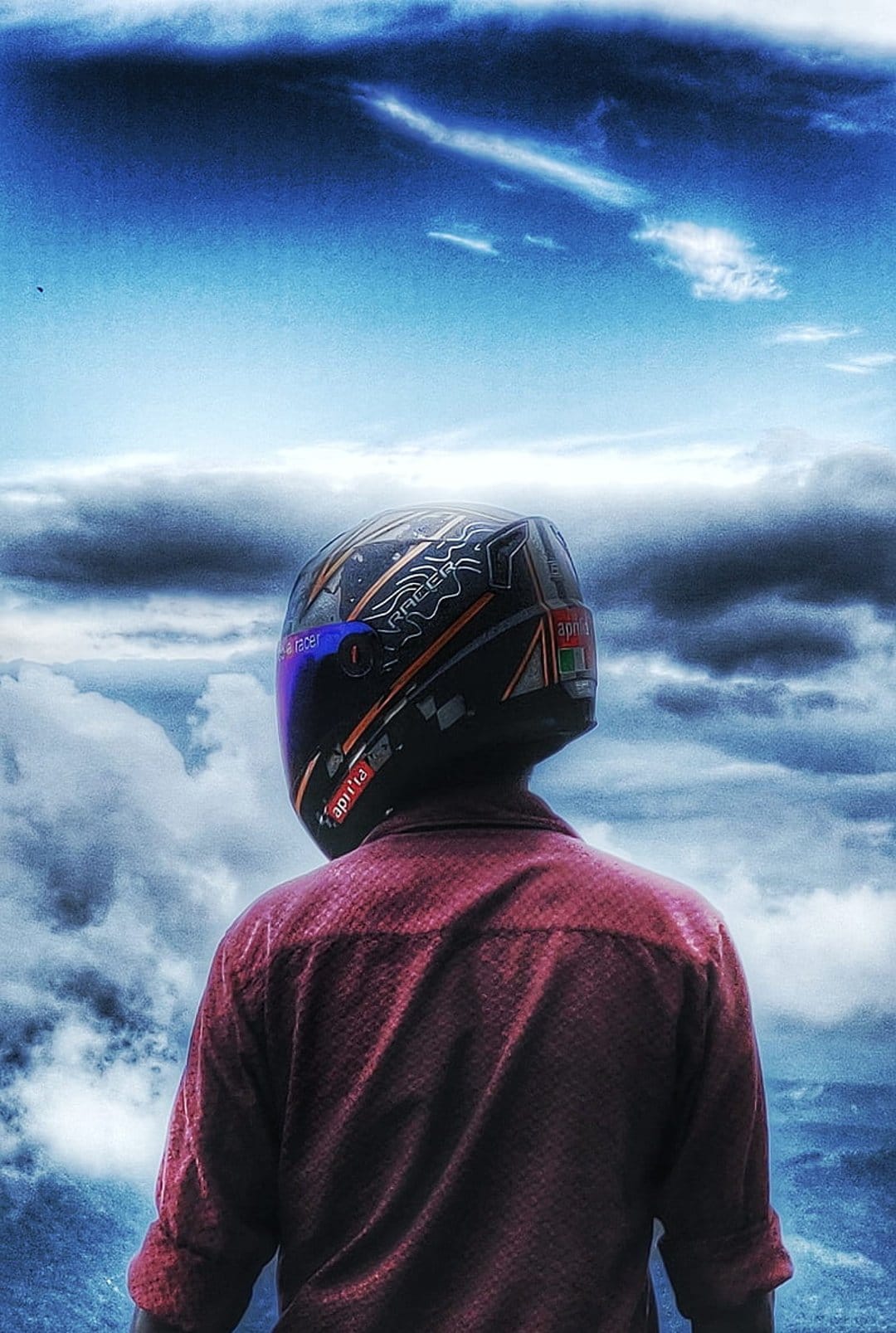 Motorcyclist in a red shirt and helmet with a purple visor looks at a cloudy blue sky with streaks of light.