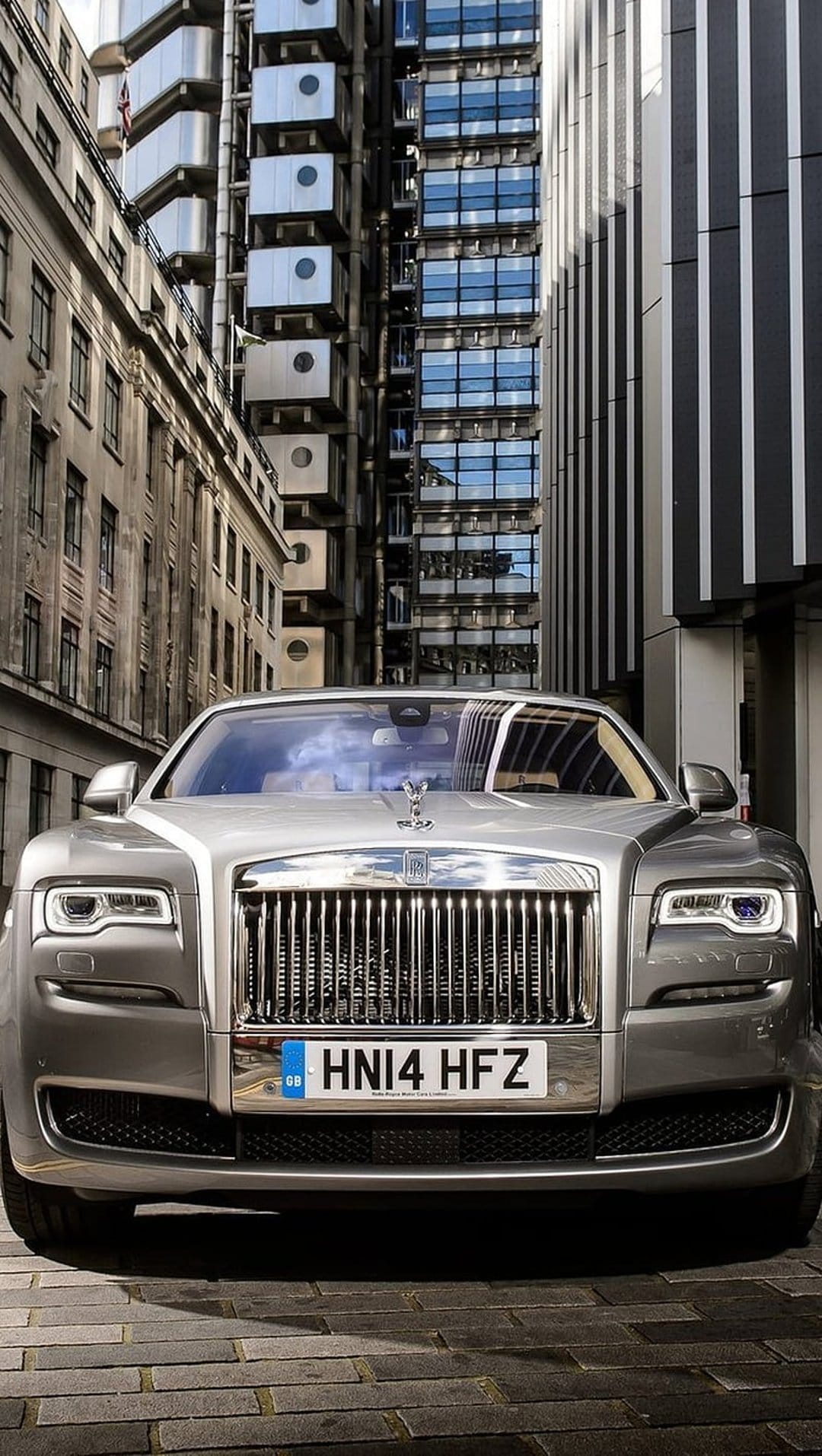 Silver Rolls-Royce Ghost parked on a cobblestone street with classic architecture and city skyscrapers.