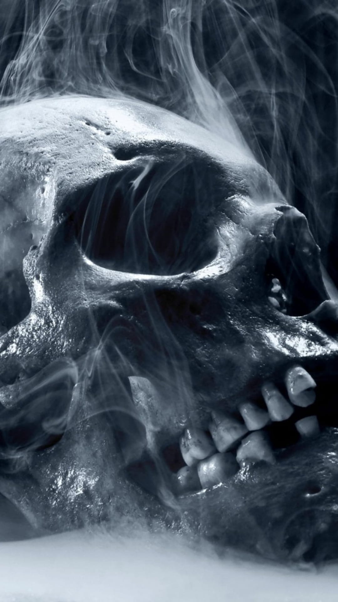 Human skull with deep eye sockets and visible teeth surrounded by wispy white smoke on a black background.