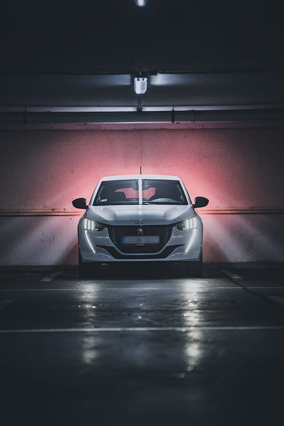 White Peugeot 208 electric car with glowing headlights in a dark garage featuring vibrant red backlighting.