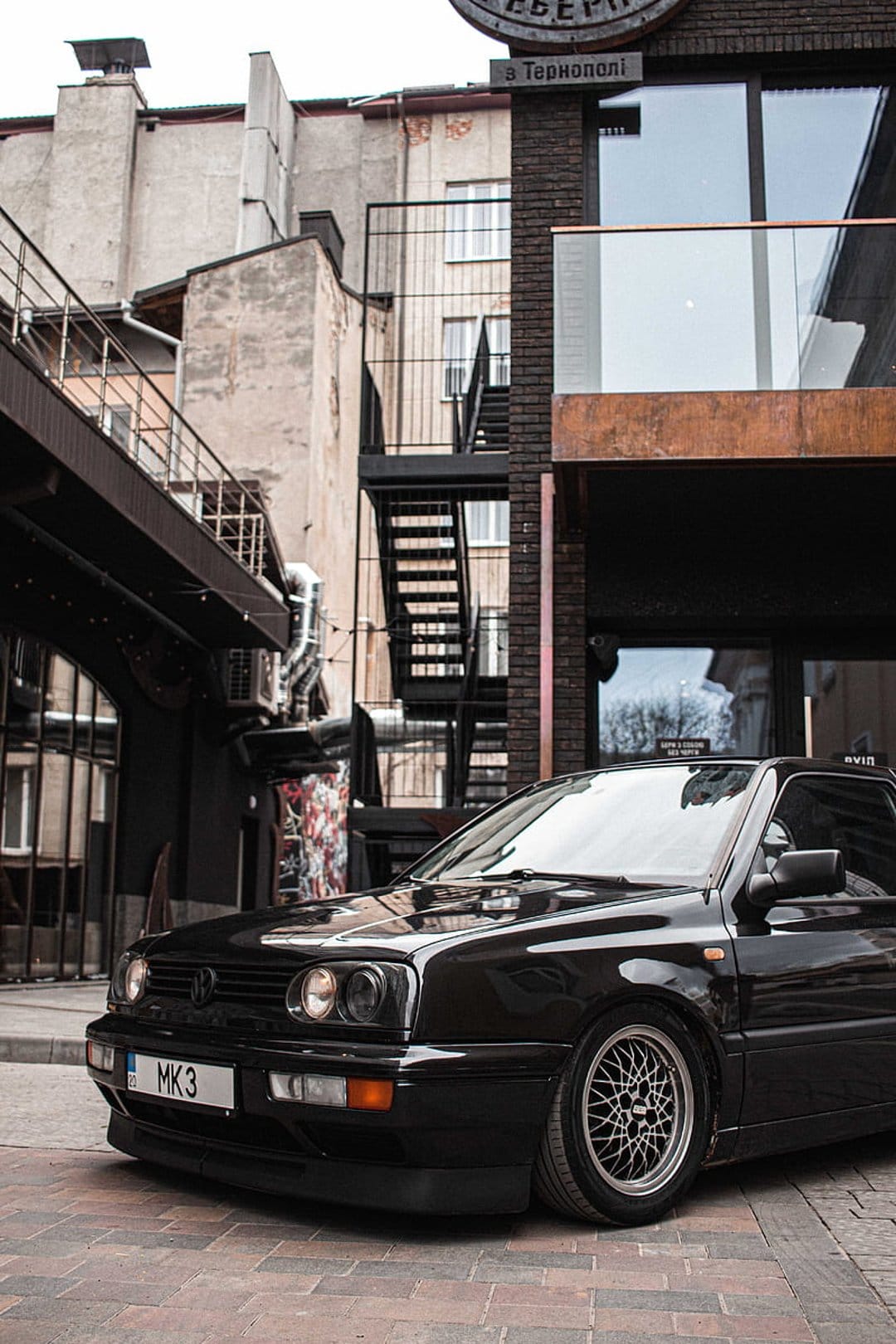 Black Volkswagen Golf MK3 with BBS wheels parked on a brick alleyway between industrial buildings with staircases.