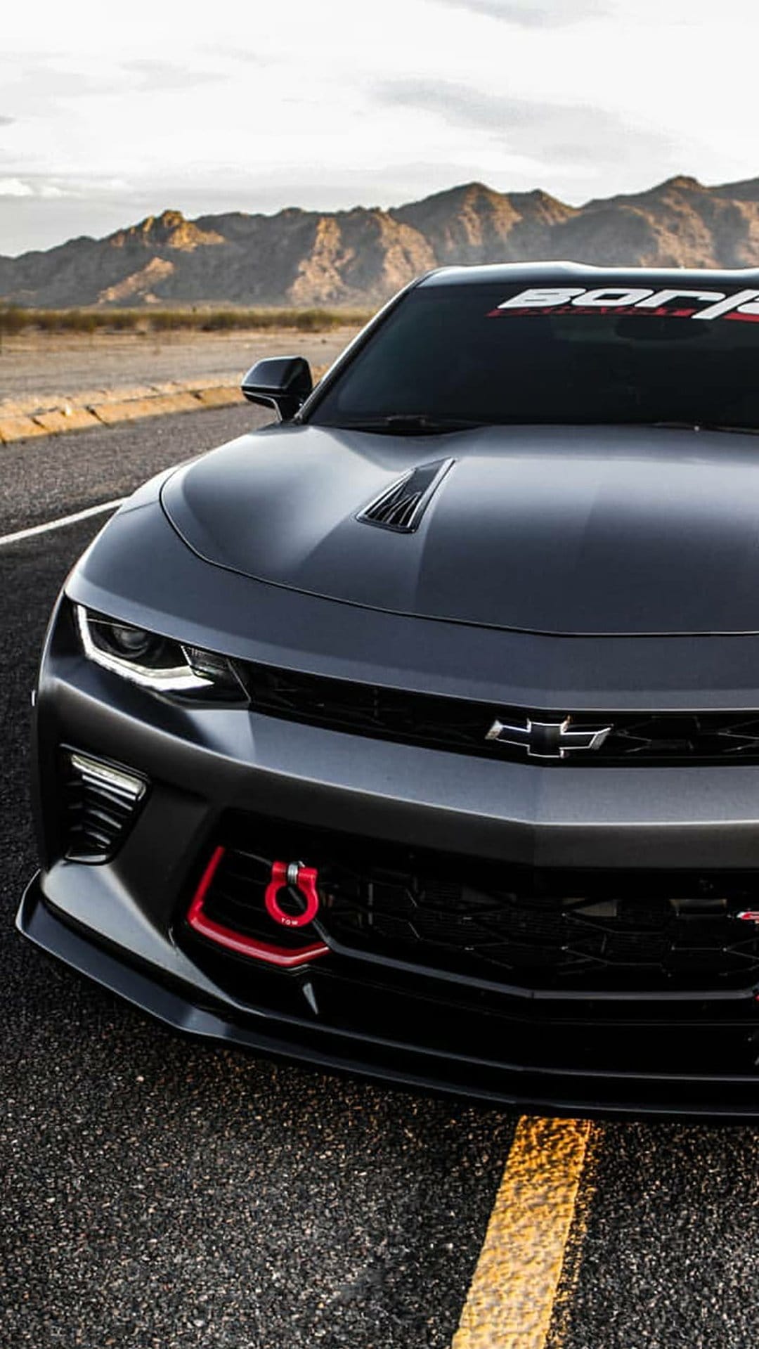 Matte black Chevrolet Camaro with a red front tow hook parked on a desert highway with mountain scenery.