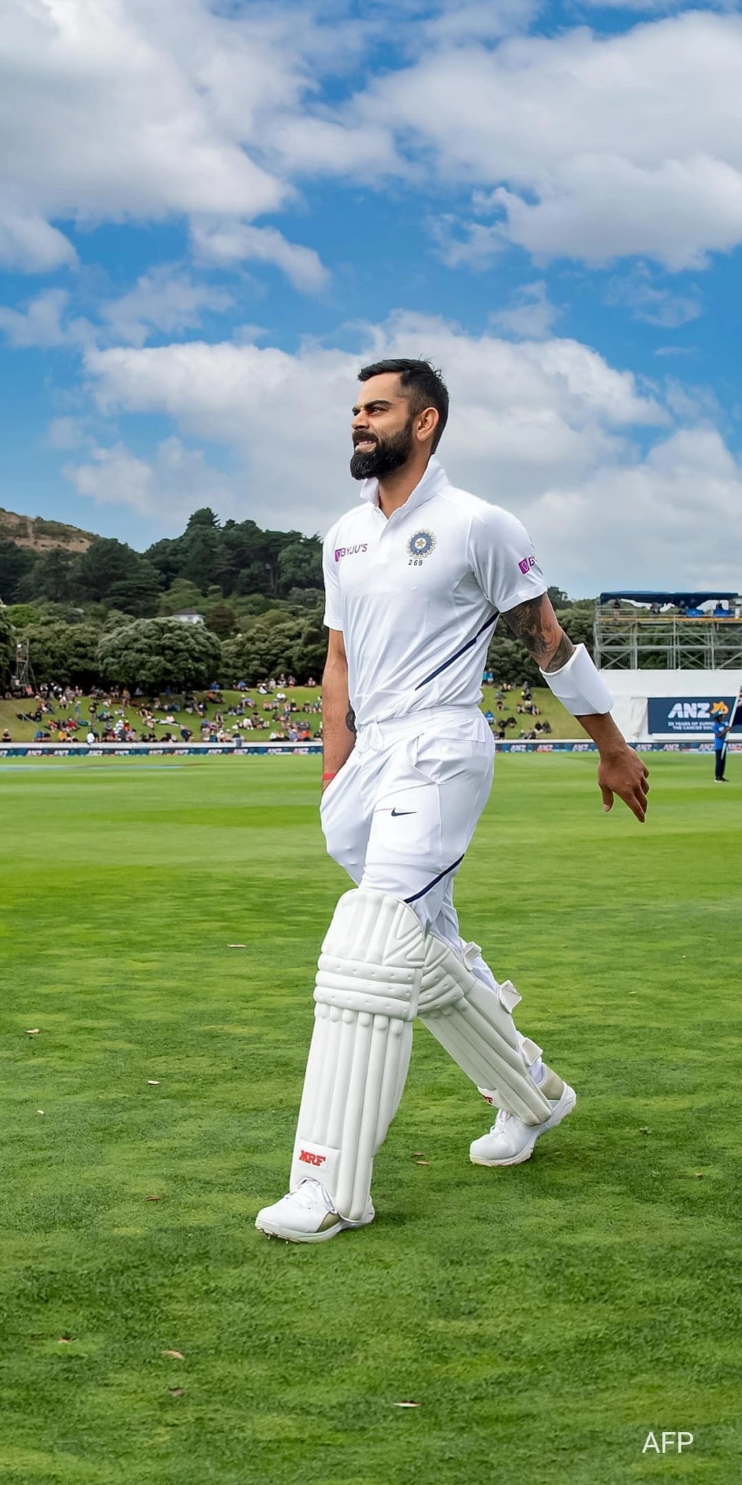 Virat Kohli in white cricket whites and pads walks across a green field under a blue sky with distant hills.
