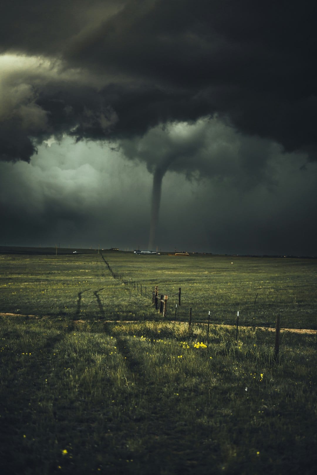 Dark funnel cloud descends onto a grassy field with a wooden fence line under a heavy, swirling storm cell.