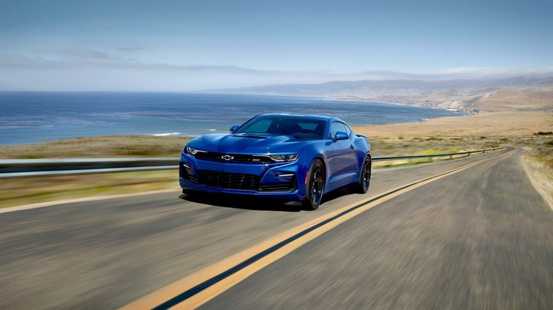 Blue Chevrolet Camaro driving on a coastal highway with ocean views and golden hills under a clear sky.