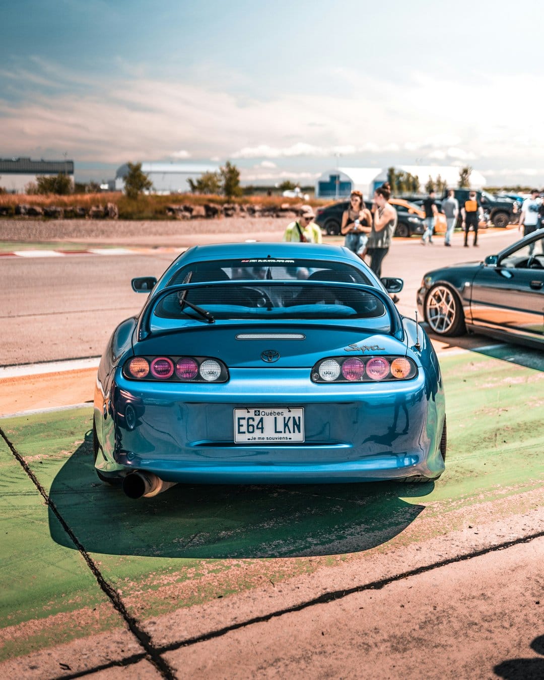 Blue Toyota Supra rear with round taillights and license plate E64 LKN parked on an asphalt race track under a blue sky.