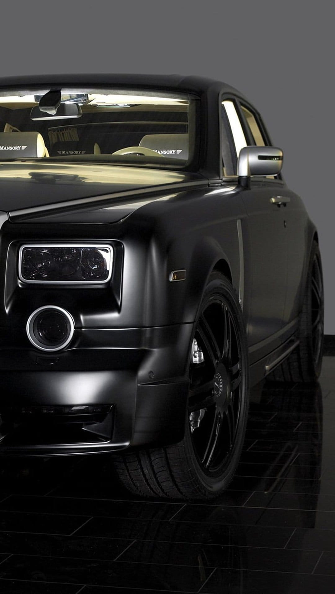 Matte black Mansory Rolls-Royce Phantom with custom wheels and headlights on a reflective studio floor.