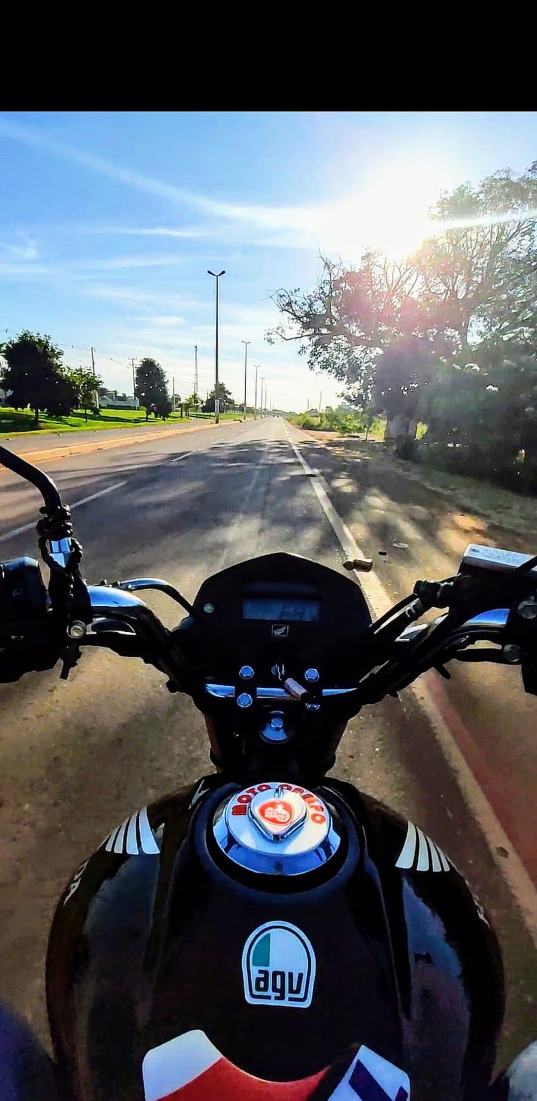 Motorcycle handlebars and a fuel tank with AGV branding overlook a sunny road lined with green trees and lamps.