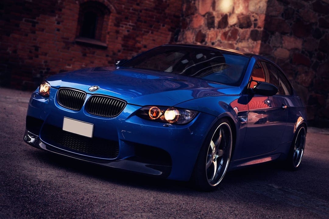 Blue BMW M3 E92 sports car with glowing orange headlights parked in front of a textured red brick wall at dusk.