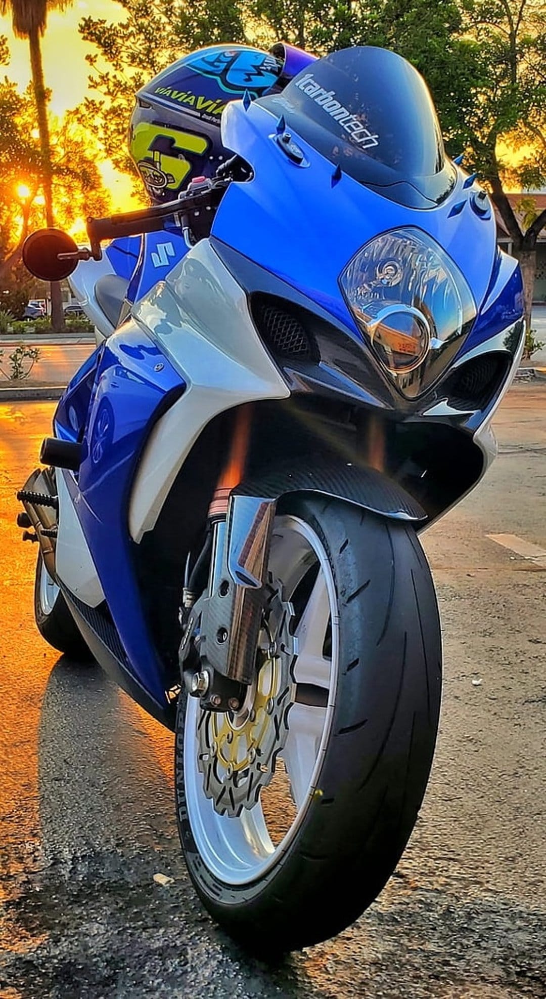 Blue and white Suzuki motorcycle with a helmet on handlebars parked on wet ground during a golden hour sunset.
