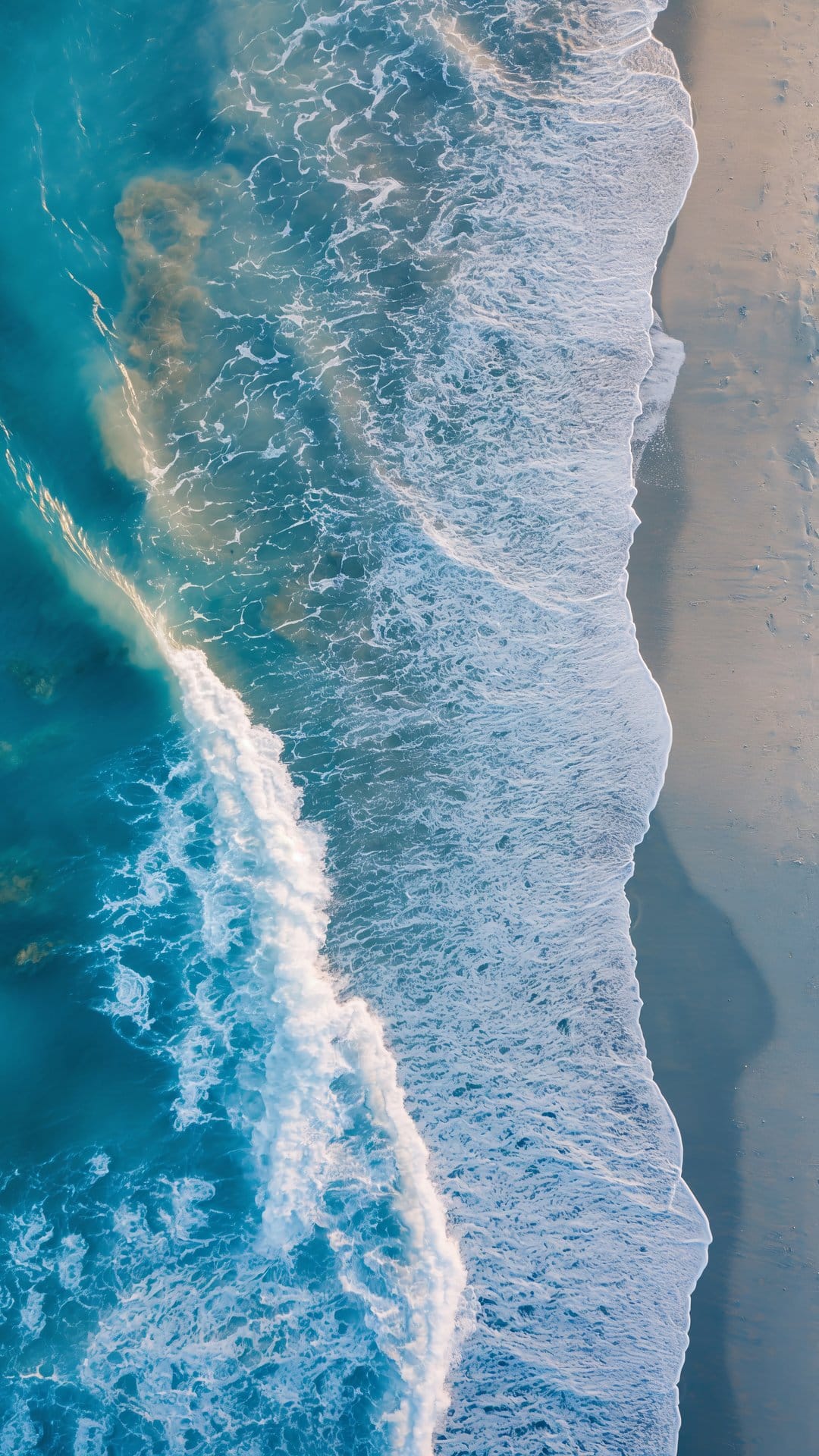 Turquoise ocean waves with white foam crests wash over a tan sandy beach in a vertical overhead drone perspective.