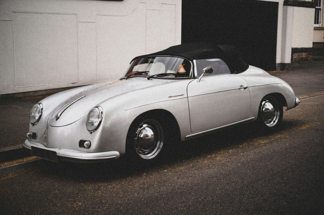 Silver Porsche 356 convertible with a black soft top parked on a street, showcasing vintage chrome trim and curves.