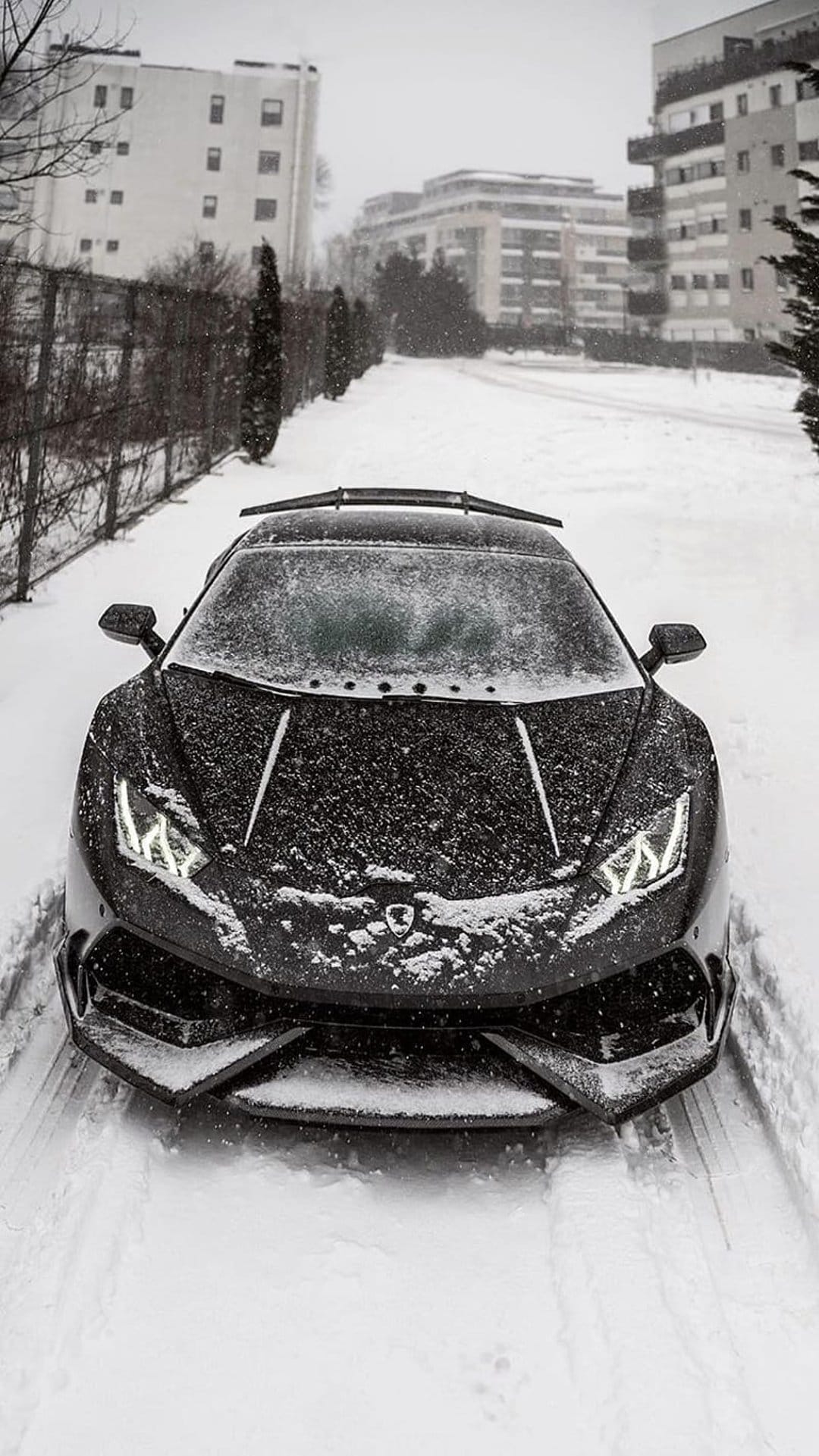 Black Lamborghini supercar with glowing headlights parked on a snowy city street during an active winter snowfall.