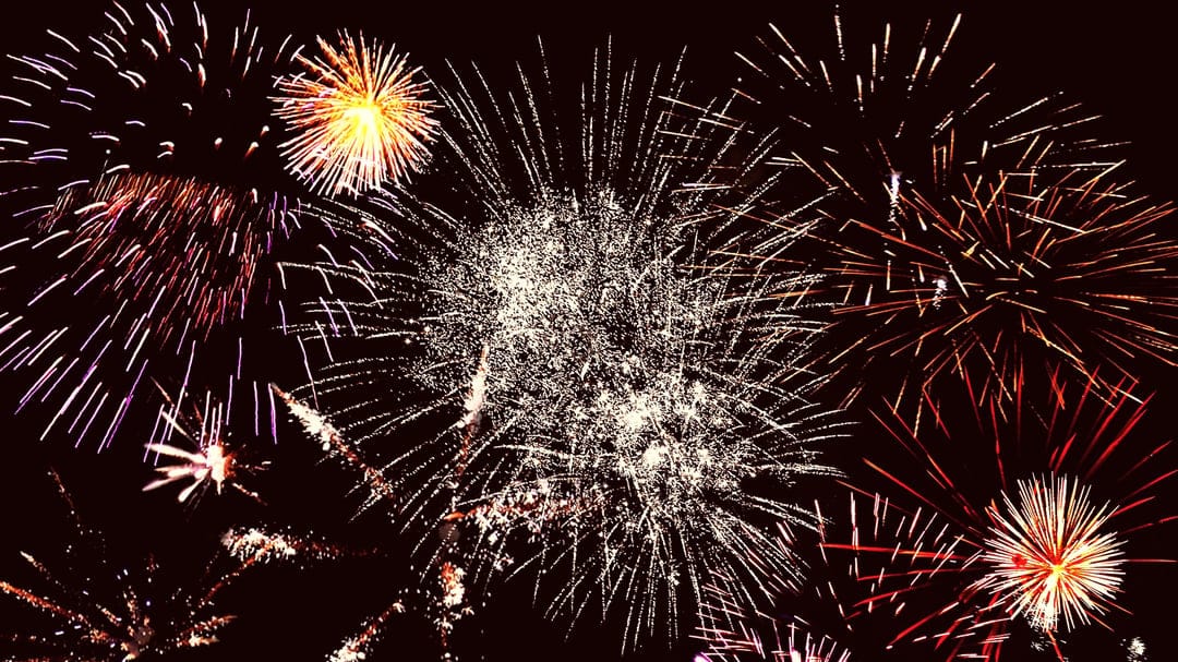 Multicolored firework bursts in pink, yellow, and white explode against a pitch-black night sky background.