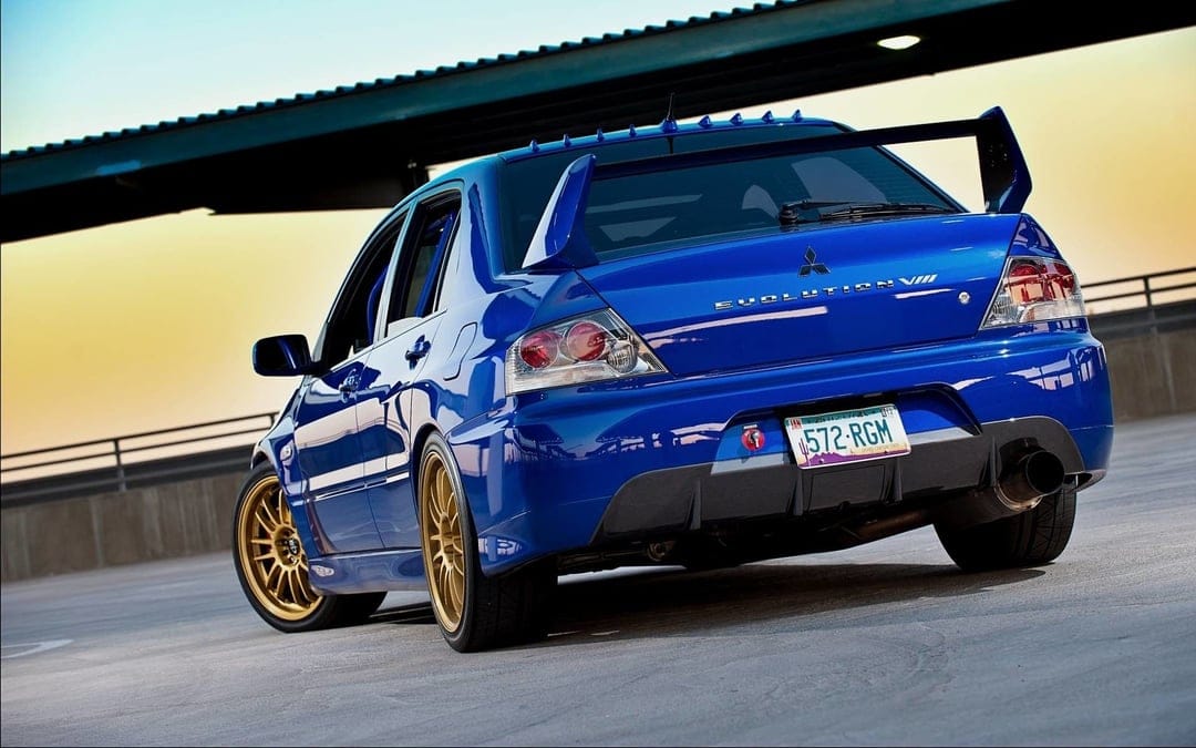 Rear view of a blue Mitsubishi Evolution VIII with gold wheels and a large wing on a rooftop at sunset.