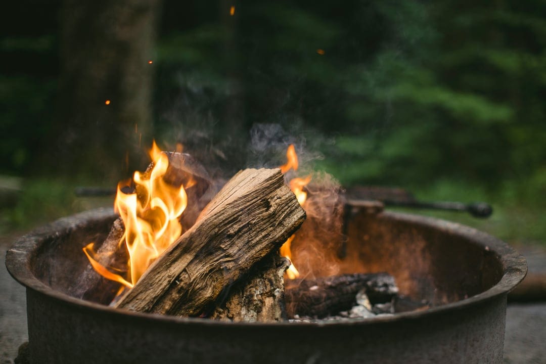 Burning logs crackle within a metal fire pit against a blurred forest background, featuring bright orange flames.
