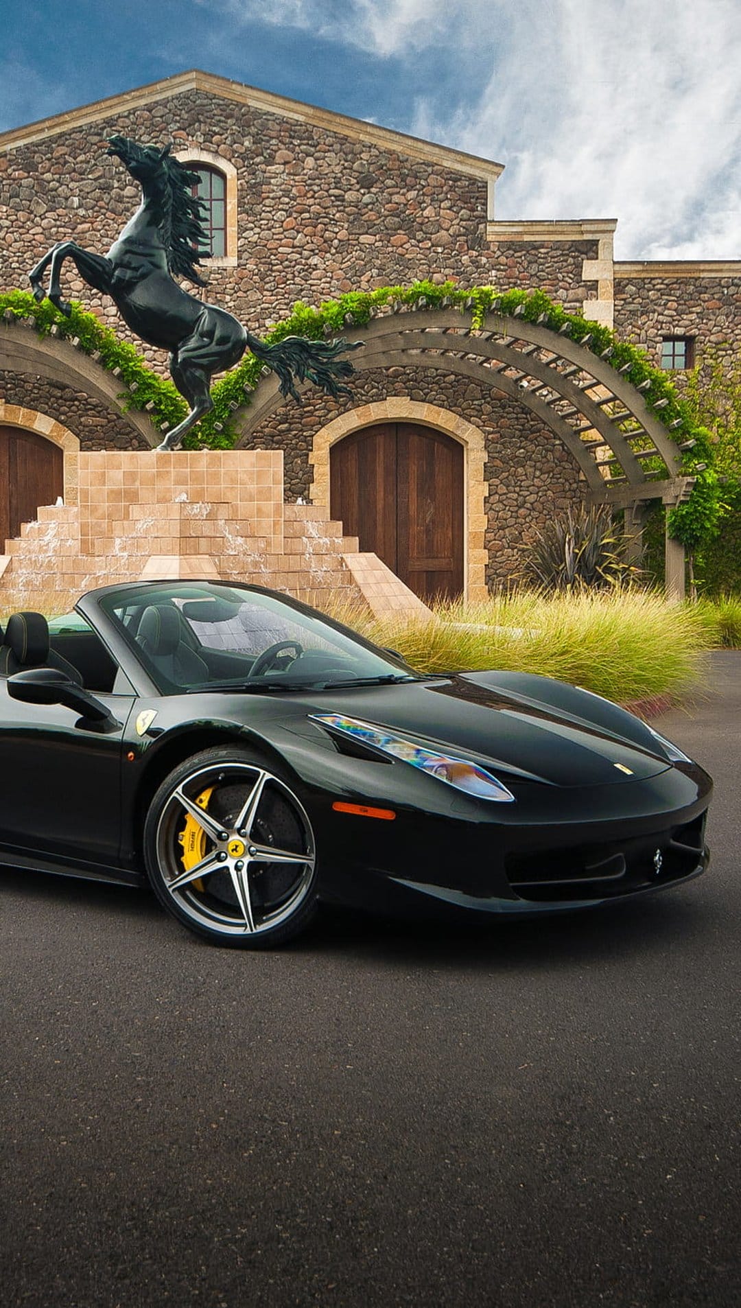 Black Ferrari convertible parked near a stone building and a galloping horse sculpture with green vines.