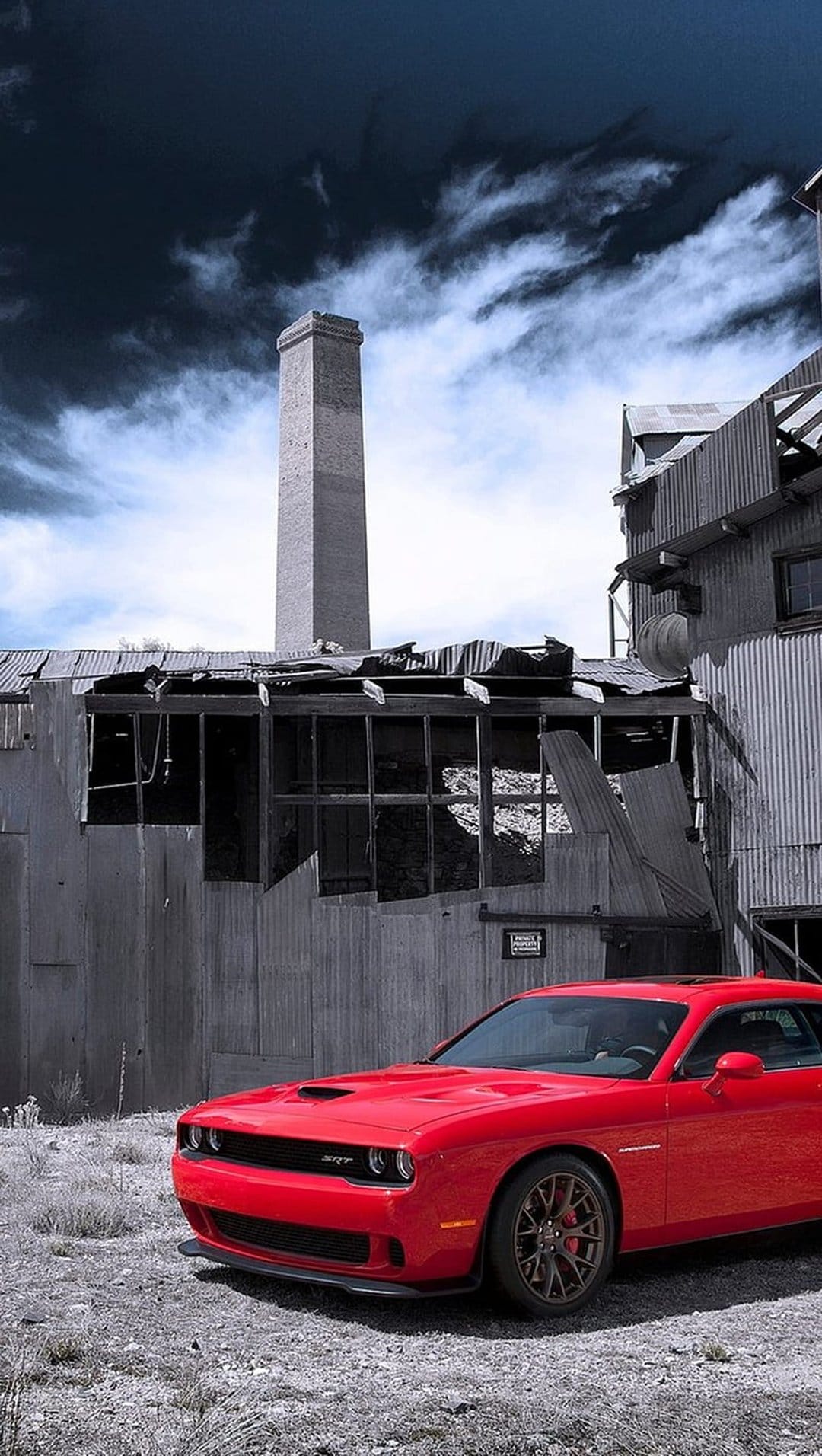 Red Dodge Challenger SRT parked on gravel in front of a tall brick chimney and abandoned industrial building.