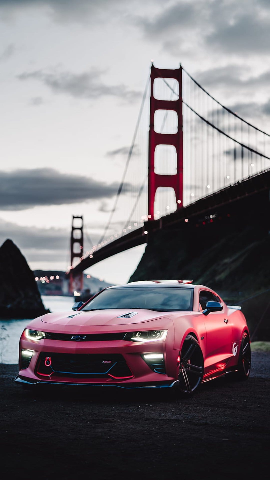 Red Chevrolet Camaro parked on dark soil with the illuminated Golden Gate Bridge under a cloudy twilight sky.
