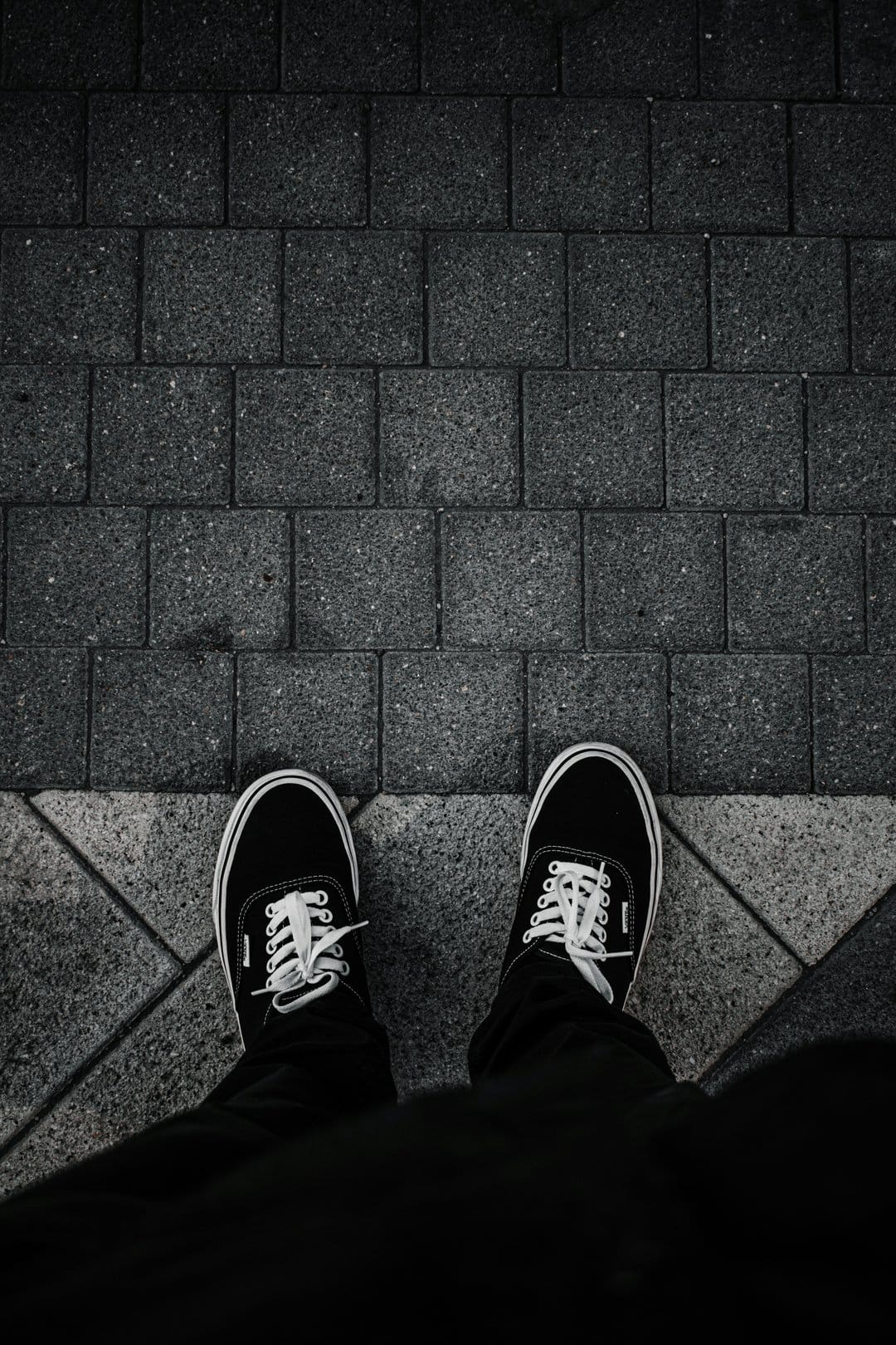 Black canvas sneakers with white laces rest on a geometric gray stone pavement in a vertical top-down view.