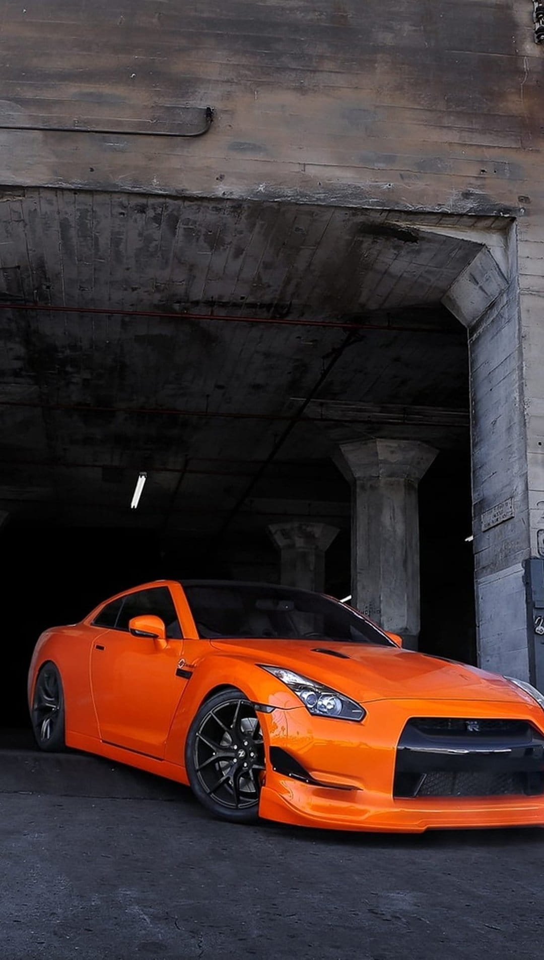 Orange Nissan GT-R parked in a dim concrete warehouse with low-key lighting and sharp reflections on the paint.