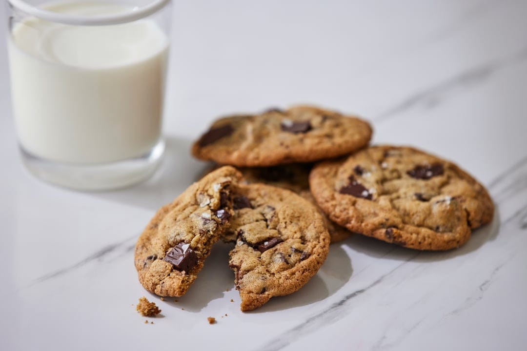 Chocolate chip cookies and a milk glass sit on white marble with scattered crumbs and melting chocolate pieces.