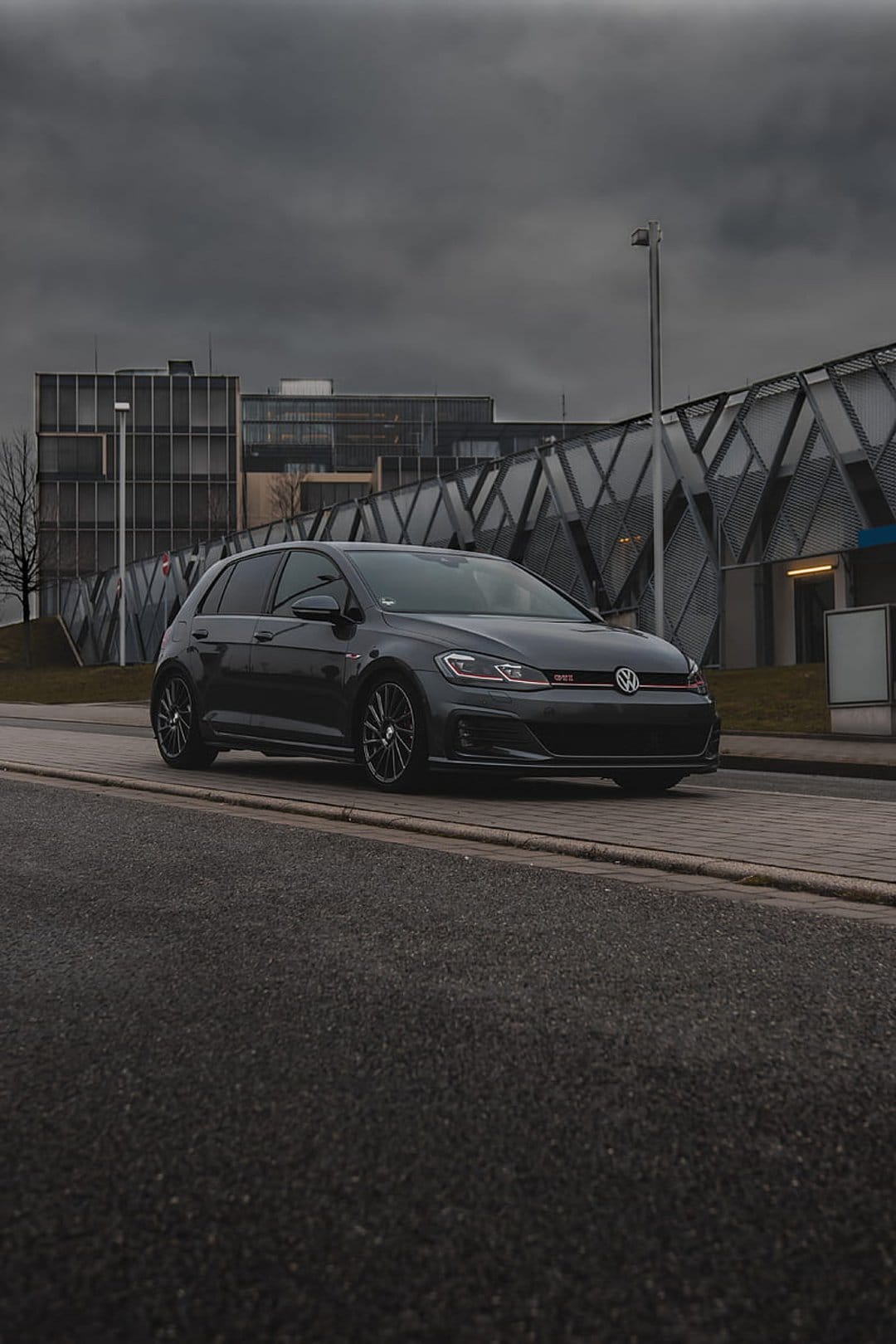 Dark gray Volkswagen Golf GTI parked on a city street near a modern building under a dramatic overcast sky.