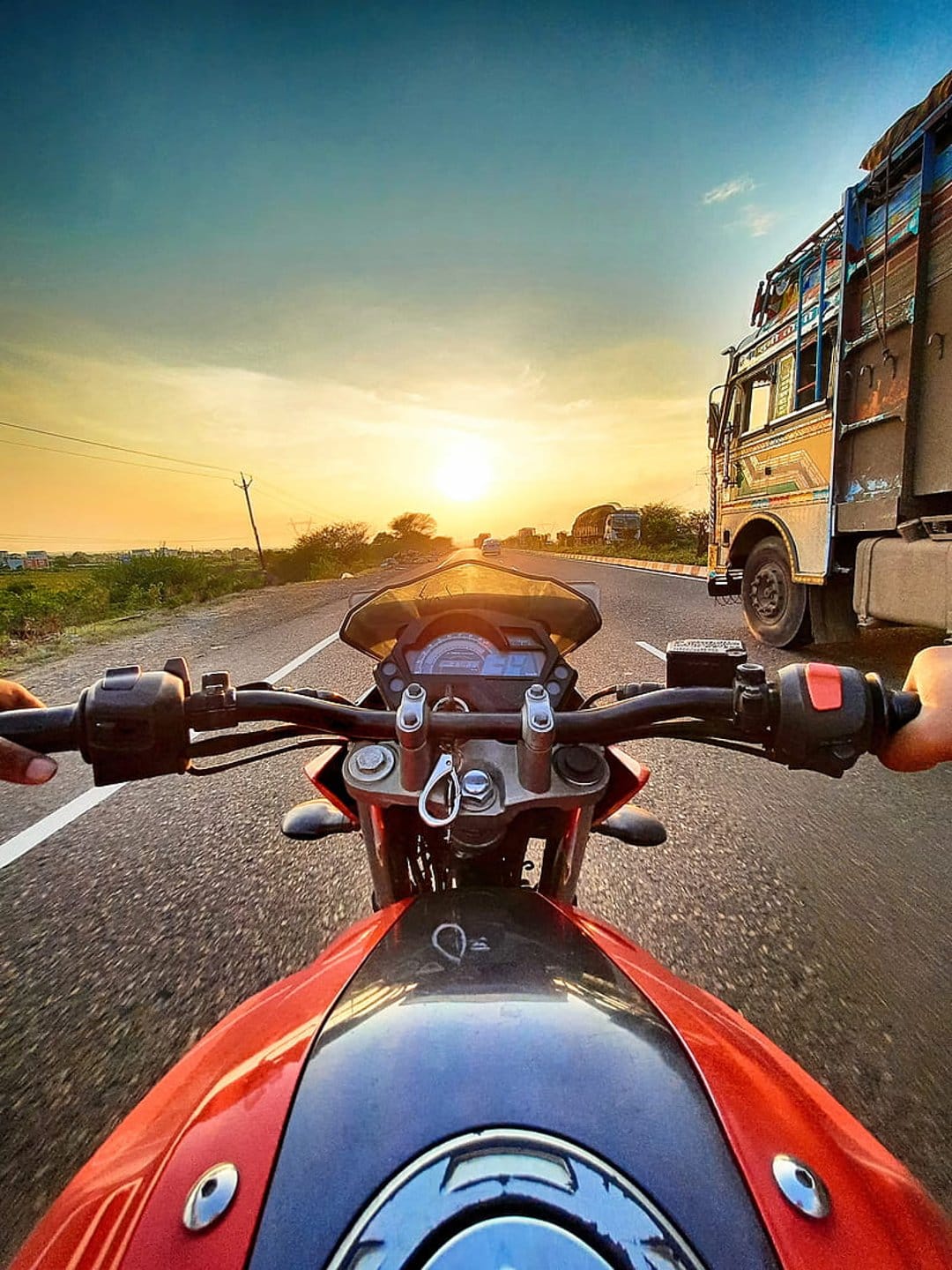 Motorcycle handlebars overlook a sunset highway with a large truck passing on the right under an orange sky.