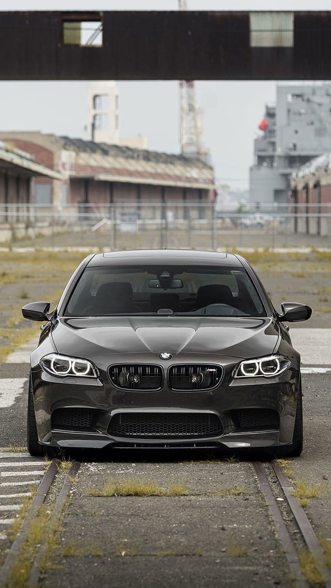 Dark gray BMW M5 sedan front view parked on weathered railroad tracks with blurred industrial cranes in the distance