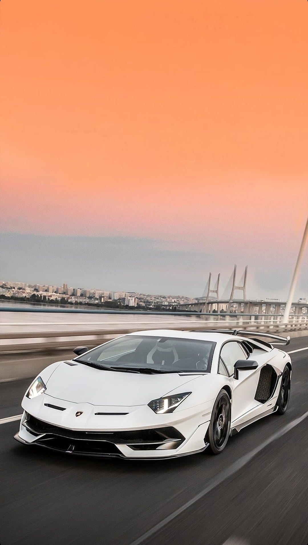 White Lamborghini Aventador on a bridge during sunset with orange sky and a city skyline in the background.